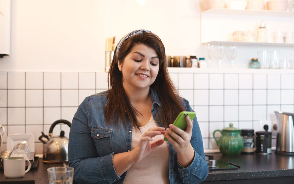 Atendimento via redes socias: Como organizar sua rotina. Mullher pluz size morena na cozinha respondendo clientes pelo celular